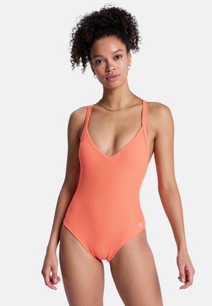 Frau mit lockigem Haar, die einen texturierten orangefarbenen Einteiler mit dünnen Trägern und einem kleinen Logo an der Hüfte trägt, vor weißem Hintergrund stehend.