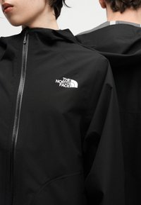 Δύο άτομα με μαύρα μπουφάν The North Face, το ένα κοιτάει μπροστά με το φερμουάρ κλειστό στον γιακά, το άλλο δείχνει την πίσω πλευρά του μπουφάν με κουκούλα.