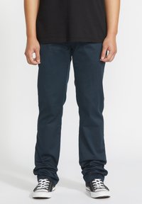 Navy blue, slim-fit pants made of smooth fabric, featuring a flat front and back pockets, paired with black sneakers and white laces.