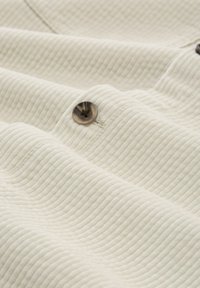 Witte, gestructureerde stof met een golfpatroon, voorzien van een donkere, ronde knoopdetail. Het materiaal lijkt zacht en licht gewatteerd.