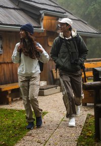 Dos personas caminan por un camino de piedras. La mujer lleva un sudadera gris, pantalones cargo beige y un gorro azul. El hombre tiene una chaqueta verde, pantalones claros y una gorra blanca.