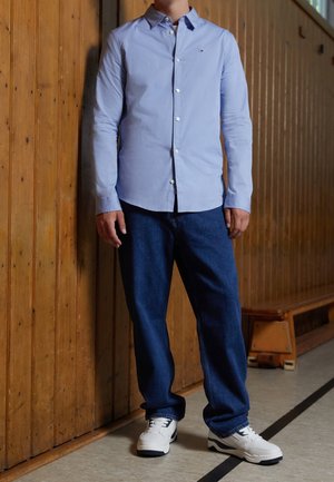 Chemise boutonnée à manches longues bleu clair associé à un pantalon en denim bleu foncé et des baskets blanches. Fond en panneaux de bois.