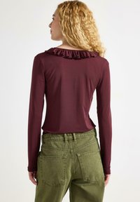 Burgunderfärgad långärmad topp med ett volangkrage, i kombination med olivgröna högmidjade byxor med bakfickor och en texturerad yta.
