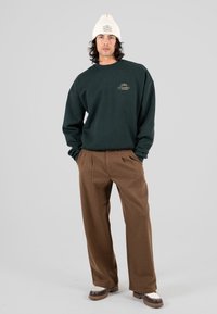 Uomo con berretto di maglia bianco, felpa verde scuro con logo, pantaloni larghi marroni e mocassini bicolore, in piedi con le mani nelle tasche.