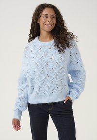 Helehelesin sinine kudum, millel on lahti istuv lõige, laineline muster, soonikkoes kaelus ja varrukad; kantud tumedate teksadega.