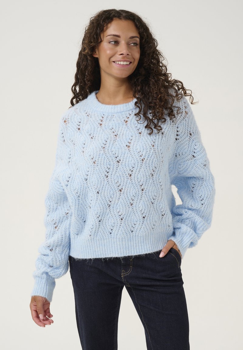 Helehelesin sinine kudum, millel on lahti istuv lõige, laineline muster, soonikkoes kaelus ja varrukad; kantud tumedate teksadega.