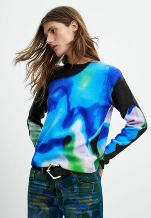 Kvinde med langt hår iført en farverig blå, grøn og sort abstrakt mønstret sweater og mørke jeans med bælte, der poserer mod en hvid baggrund.