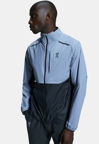Helleblaue und schwarze Jacke, entworfen mit einem Stehkragen, Reißverschluss und elastischen Bündchen. Verfügt über ein Logo und kontrastierende Farbpaneele.