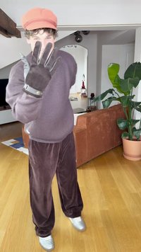 Barna kordbársony nadrág, texturált lila pulóver, szürke kesztyűk díszgombbal, és narancssárga sapka. Beltéri környezet növénnyel és kanapéval.