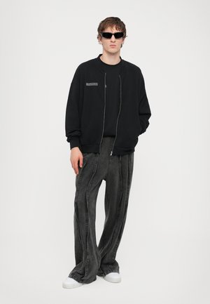 Svart zip-up bomberjakke over en svart t-skjorte, paret med løstsittende grå denimbukser. Modellen har på seg mørke solbriller og hvite sneakers.