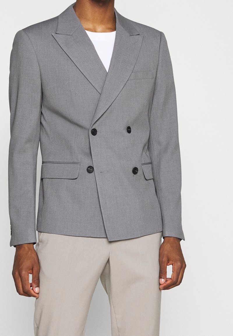 Blazer gris à double boutonnage avec revers, poche poitrine simple et boutons noirs. Porté avec une chemise blanche et un pantalon de couleur claire.