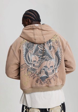 Homme aux cheveux tressés portant un sweat à capuche marron avec des crânes ailés, des roses et un motif "SEATTLE" dans le dos, sur une chemise blanche et un pantalon à motifs.