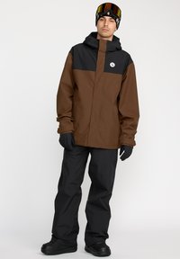 Brun og sort skijakke med et logo-patch, parret med sorte bukser og handsker. Har hætte og en afslappet pasform. Snebriller på panden.