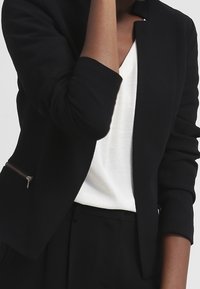 Blazer noir à la coupe ajustée, avec un col structuré et des détails zippés sur les manches. Porté sur un haut blanc.