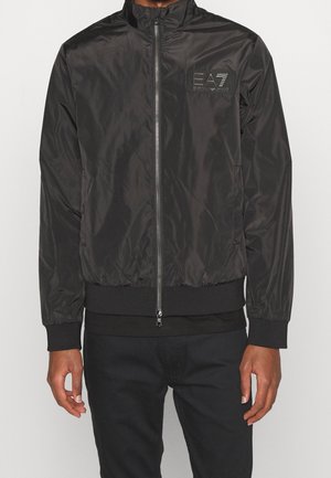 Blouson bomber noir zippé avec logo EA7 Emporio Armani sur la poitrine gauche, porté par une personne en pantalon noir.