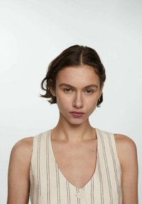 Jongere met kort bruin haar, gekleed in een mouwloos beige top met verticale strepen, tegen een effen witte achtergrond.