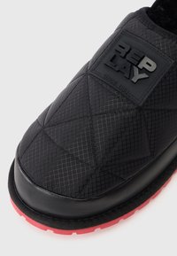 Zapato negro acolchado tipo slip-on con suela de goma que presenta un acento rojo, tejido texturizado y un prominente parche con el logo negro.