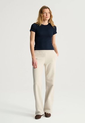 Kobieta stojąca z rękami w kieszeniach, ubrana w granatową koszulkę z krótkim rękawem, beżowe spodnie i brązowe buty na jednolitym jasnym tle.