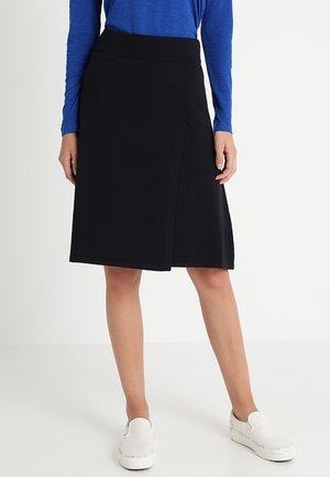 Femme portant une jupe portefeuille noire longueur genou, un haut bleu à manches longues et des baskets blanches sans lacets, debout sur un fond blanc.