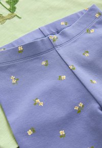 Nahaufnahme eines violetten Stoffs mit kleinen gelben Blumenmustern und sichtbaren Nähten auf einem darunter liegenden, gefalteten grünen Kleidungsstück.
