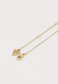 Collier en chaîne dorée avec un pendentif triangulaire portant l'inscription "GUESS" gravée et une fermeture à mousqueton. Fini lisse et poli.