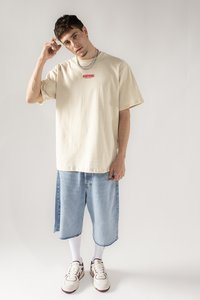 Beige Oversized-T-Shirt mit rotem Text, ausgebleichte blaue Denim-Shorts mit rohem Saum, weiße Socken und klobige weiße Sneakers mit burgunderfarbenen Akzenten.