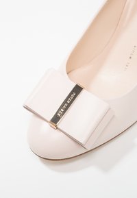 Botte plate en cuir rose pâle avec un bout arrondi, ornée d'un grand nœud et d'une quincaillerie dorée accentuant le centre. Texture lisse sur l'ensemble.