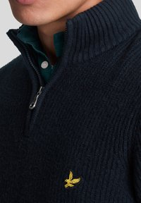 Suéter de punto azul marino con un alto cuello y media cremallera. Presenta un sutil patrón texturizado y un pequeño emblema de águila amarilla en el pecho.