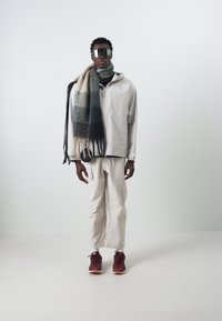 Lätt, beige jacka med ståkrage, tillsammans med lösa krämfärgade byxor. En flerfärgad scarf hänger över halsen, kompletterad med mörka sneakers.