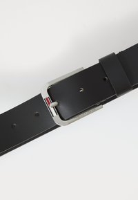 Cinturón de cuero negro con hebilla de plata que presenta franjas en rojo, blanco y negro, y con "Tommy Jeans" grabado en la barra de la hebilla.