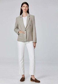 Veste légère gris clair avec deux poches avant, fermeture par boutons, et un motif brodé partiel. Associée à un pantalon blanc et des mocassins marron.