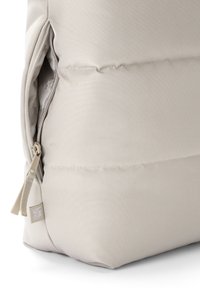 Colțul unei genți din material bej cu buzunar cu fermoar parțial deschis, care arată interiorul cu model și o mică etichetă atârnată de fermoarul buzunarului.