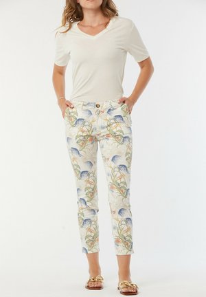 Pantalon à motif floral blanc avec des accents bleus et verts, associé à un t-shirt beige clair en V. Le modèle se tient avec les mains dans les poches.