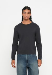 Jonge man met gevlochten haar, gekleed in een zwart shirt met lange mouwen en blauwe jeans, staande tegen een effen witte achtergrond.