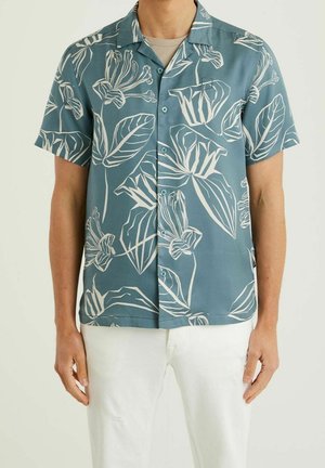 Mand iført blå kortærmet skjorte med hvide blomsterprint og hvide bukser, stående mod en ensfarvet baggrund.
