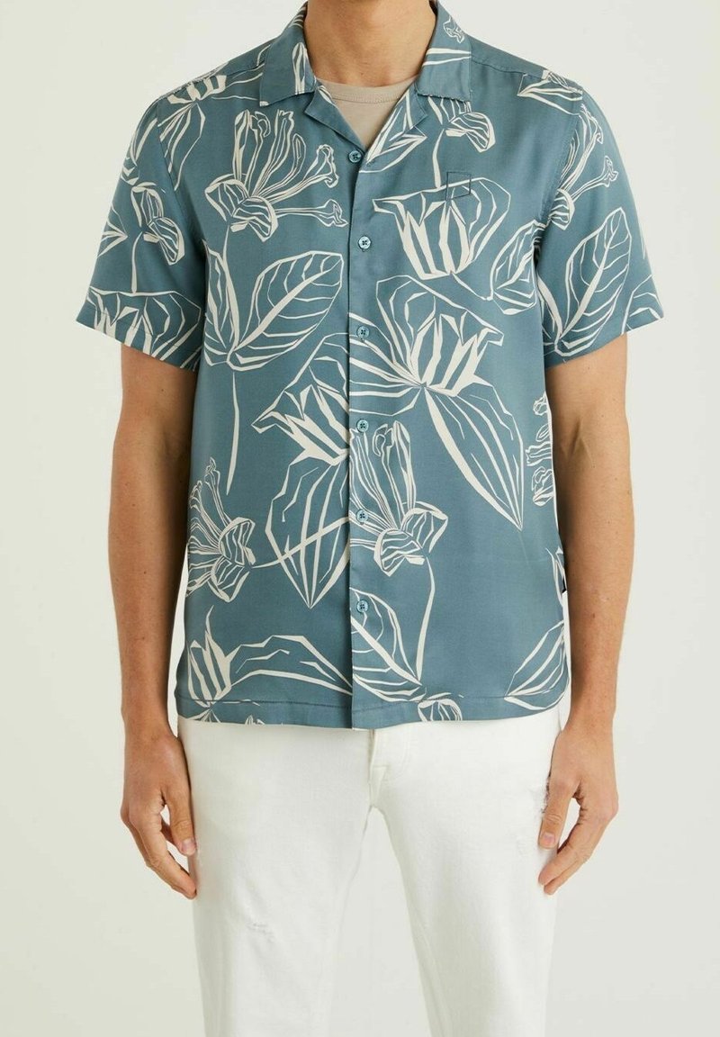 Homme portant une chemise bleue à manches courtes avec un imprimé floral blanc et un pantalon blanc, debout devant un fond uni.
