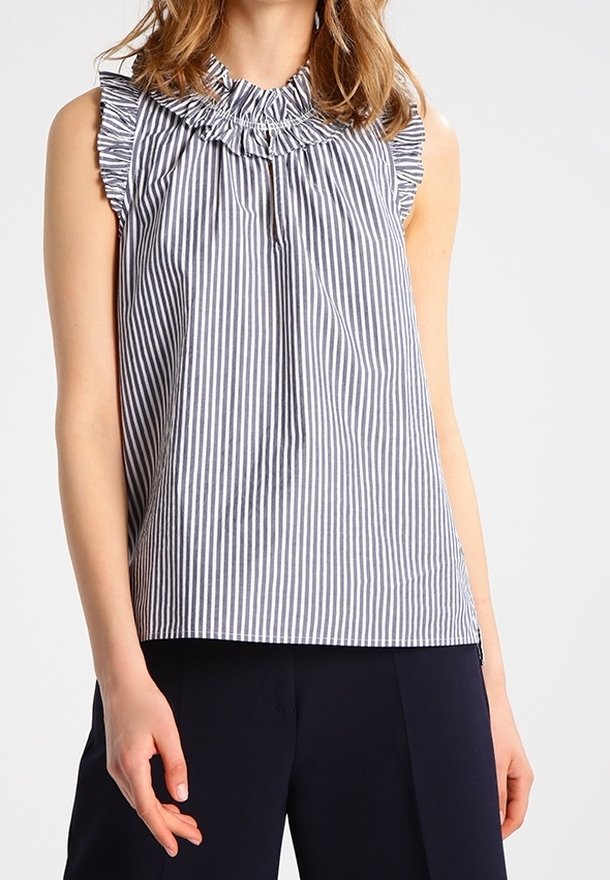 Blouse sans manches rayée verticalement bleu et blanc avec col et manches à volants portée avec un pantalon foncé.