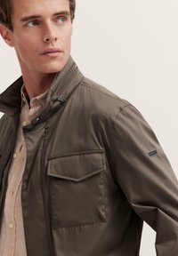 Leichte olivgrüne Jacke mit hohem Kragen, Reißverschluss vorne und einer Brusttasche mit Klappe. Enthält ein markantes Detail am Ärmel.
