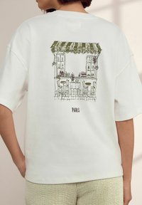 Vit bomulls-T-shirt med en grön illustrerad kafé-scen på baksidan med texten "PARIS" nedanför. Korta ärmar, avslappnad passform.