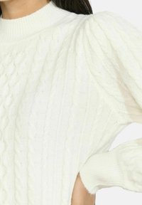 Maglione bianco lavorato a maglia con collo alto, caratterizzato da motivi a trecce e maniche a sbuffo. La texture appare morbida e accogliente.
