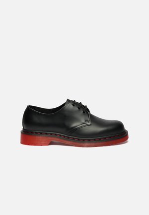 Schwarzer Lederschnürschuh mit dicker roter Sohle und roter Naht auf weißem Hintergrund, Seitenansicht.