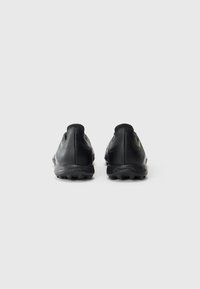 Une paire de crampons de football noirs, à tige basse, avec une surface texturée et de petits crampons sur les semelles, vue de l'arrière sur un fond uni.
