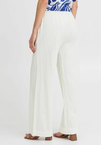 Pantalon large en lin blanc avec une taille élastique, présentant une finition texturée et une coupe décontractée. Sandales à talons carrés brunes visibles.
