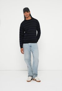 Pull marinière, coupe décontractée, porté avec un jean bleu clair et des baskets blanches. Une casquette noire finit la tenue décontractée.