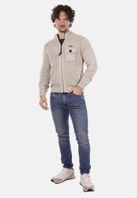 Maglione beige lavorato a maglia con zip e colletto alto, dotato di tasca frontale e bottone nero. Abbinato a jeans blu e sneakers bianche.