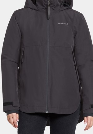 Veste noire à capuche Didriksons avec fermeture éclair frontale, poignets en Velcro et poches latérales portée avec un pantalon noir.