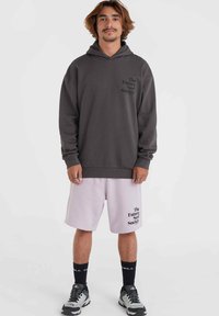 Sudadera gris con logo negro en el pecho, combinada con pantalones cortos rosa claro que presentan un logo similar. Calcetines negros y zapatillas deportivas grises completan el atuendo.