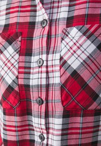 Chemise en flanelle à carreaux rouges et blancs avec des accents noirs, comportant deux poches poitrine et des boutons noirs sur le devant. Texture de tissu douce.