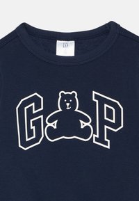 T-shirt pour bébé bleu marine avec un col rond, présentant une silhouette blanche d'ours et les lettres "GAP" en gras, de couleur blanche contrastante.