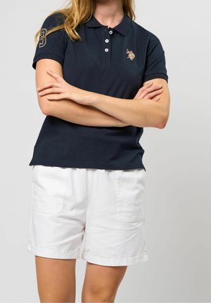 Kobieta ubrana w granatową koszulkę polo z haftowanym logo i białe szorty z podwiniętym brzegiem, z założonymi ramionami na jednolitym tle.